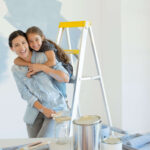 Mother with her daughter on back laughing as they paint a room in front of a ladder